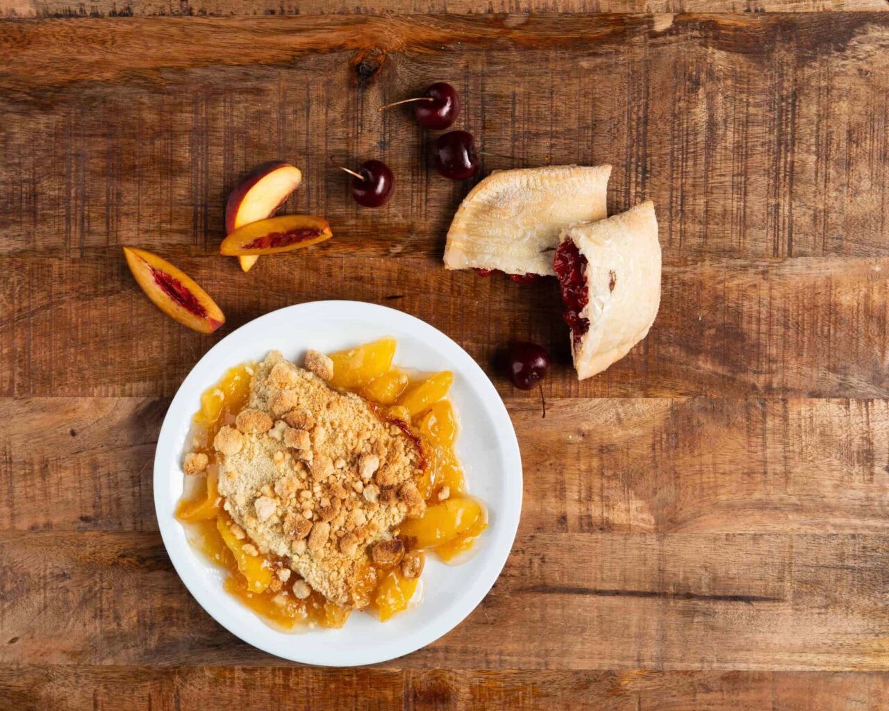 Peach crumble on a plate next to a slice of cherry pie and fresh cherries, showcasing store prepared desserts.